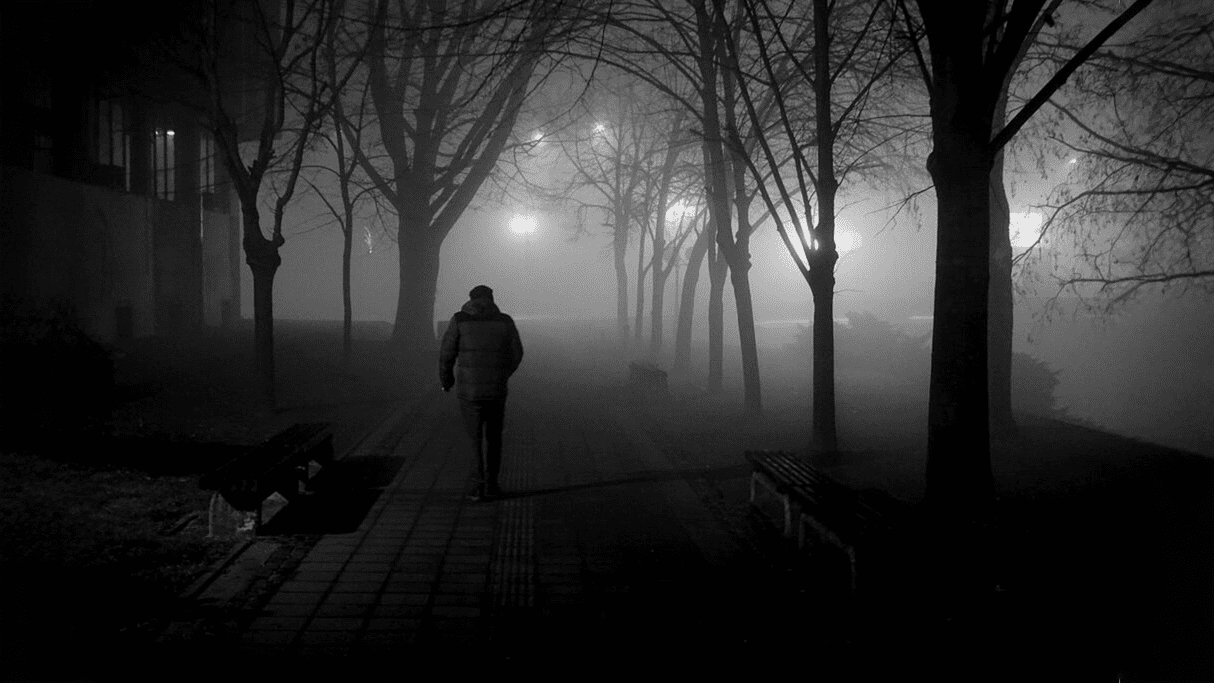 A lone figure walking away down a paved path through a dark, foggy park, illuminated by the glow of streetlights in the distance.