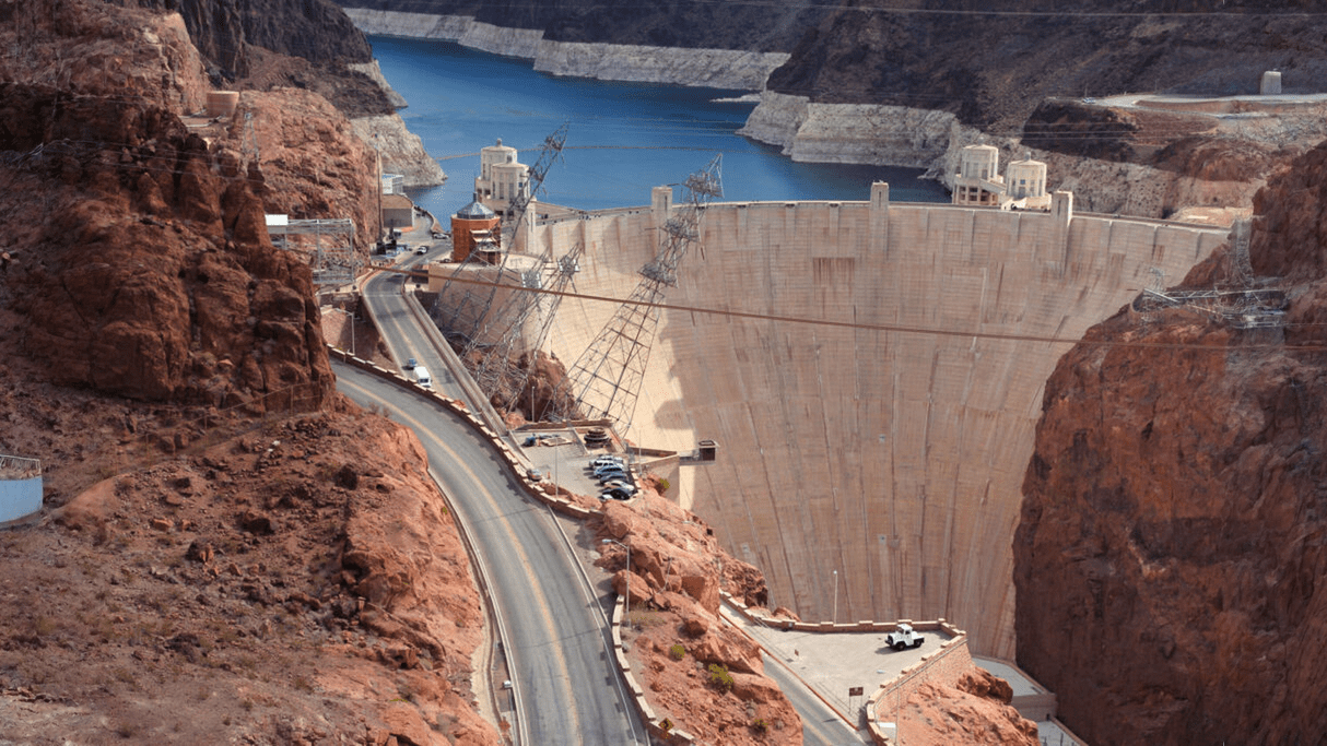 High-angle aerial view of the Hoover Dam and the Colorado River, showing the massive curved concrete wall and electrical towers against the canyon rocks.
