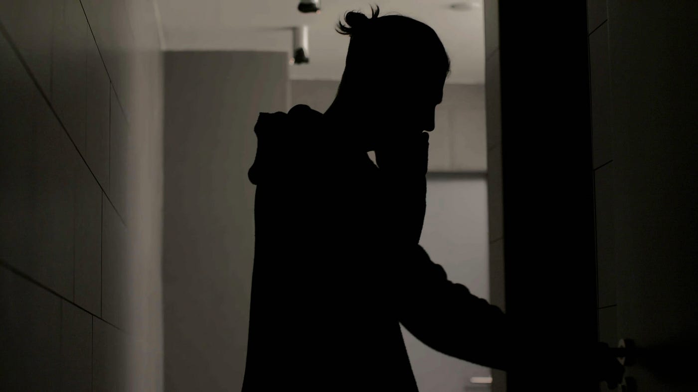 A student stands in silhouette in a dim school hallway, with one hand raised to their face.