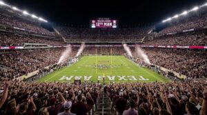 Panoramic view of a packed Kyle Field at Texas A&M at night with the Jumbotron displaying "12TH MAN" and the crowd standing.